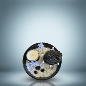 An elegant decorative arrangement on a granite countertop, featuring a white candle wrapped in a black ribbon, a large black crystal in a crown-shaped holder, smaller black stones on iridescent material, and purple fabric flowers