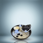 A decorative gift set featuring a candle, a large raw black tourmaline stone, several small tumbled black tourmaline stones, and decorative elements arranged in a black bowl filled with translucent snowflakes