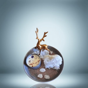 A decorative setup featuring a golden reindeer figurine, a candle with rose petals and crystals, a large clear quartz cluster, and three smaller quartz stones arranged on a gray plate with a textured mat