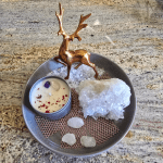 "A decorative gift set featuring a golden reindeer figurine, a small container of white cream or wax with red and purple elements on top, a large clear quartz crystal, and several smaller clear quartz stones, all arranged on a round, dark-colored plate with a textured mat underneath some of the elements.