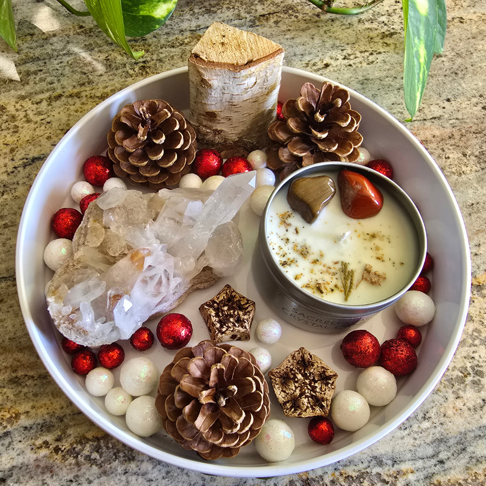 A decorative arrangement featuring pinecones, red and white glittery balls, a piece of wood, a cluster of clear quartz crystals, and a candle in a metal tin with two polished stones embedded in it, all displayed on a granite countertop