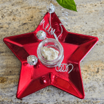 A decorative red star-shaped ornament with a small container of muscovite in the center, adorned with clear beads and silver wire spirals