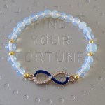 Translucent opalescent bead bracelet with small gold spacer beads and an infinity symbol charm, half encrusted with rhinestones and half dark blue.