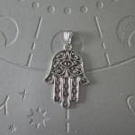 A silver-finished brass pendant shaped like the Hamsa hand, with intricate filigree designs, measuring 2 inches in size.