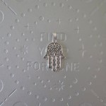 A silver-finished brass pendant shaped like the Hamsa hand, with intricate filigree designs, measuring 2 inches in size.