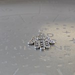 A silver-finished brass pendant shaped like the Hamsa hand, with intricate filigree designs, measuring 2 inches in size.