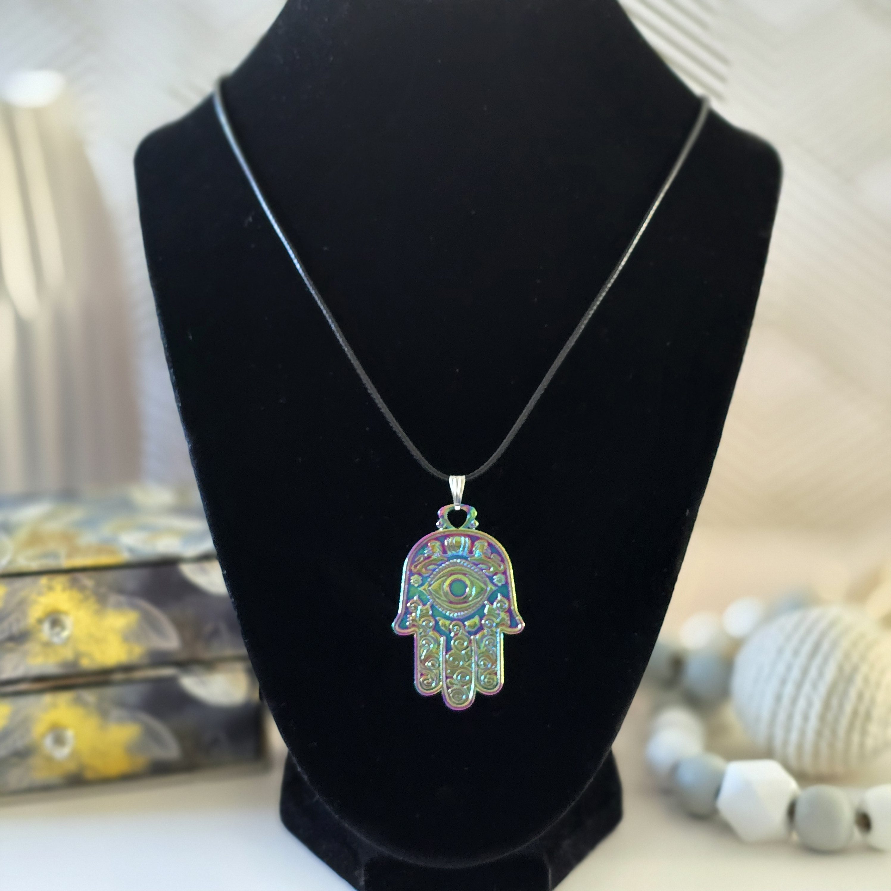 A necklace featuring a Hamsa hand pendant with an iridescent finish, displaying intricate patterns and vibrant colors, on a 22-inch black cord.