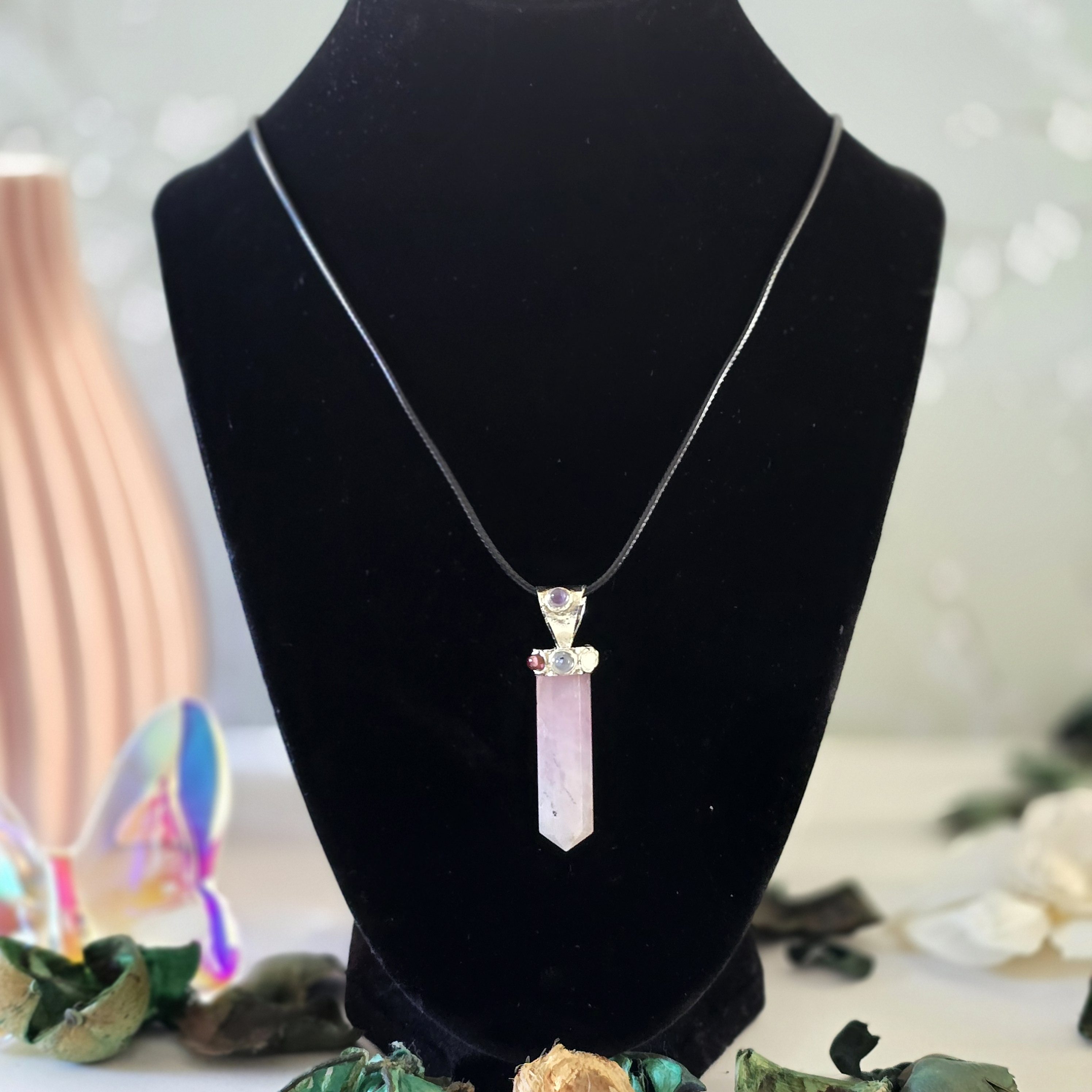 A rose quartz pendant paired with tiny chakra stones, hanging from a black cord, displayed on a black velvet stand with decorative elements in the background.