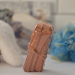 A Palo Santo fizz bath bomb wrapped in clear plastic, with a reddish-brown color and a textured surface, displayed with a blue bath sponge in the background.