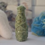 A sage purity fizz bath bomb wrapped in clear plastic, with a greenish hue and a textured surface, displayed with a blue bath sponge in the background.